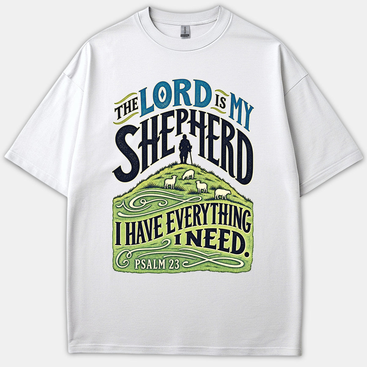 A white Christian T-shirt featuring decorative typography reading 'The Lord Is My Shepherd' above an illustration of a shepherd and sheep on a green hill with Psalm 23.