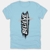 A light blue t-shirt featuring a Christian graphic design with a gray cross over a distressed black background and the word BELIEVE in bold white text.