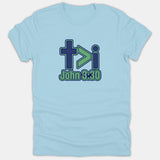 A light blue T-shirt featuring a Christian graphic with a cross, greater-than symbol, and letter 'i' above the Bible reference John 3:30 printed in navy and green.