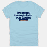 A light blue t-shirt featuring the phrase 'by grace. through faith. not works.' in bold navy text with the scripture reference 'EPHESIANS 2:8-9' in orange.