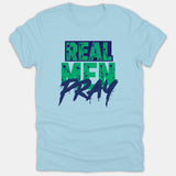 A light blue t-shirt featuring a bold graphic design with the phrase REAL MEN PRAY printed in green and navy distressed splatter lettering.