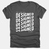 A dark gray Christian t-shirt featuring bold white text reading THIS T-SHIRT WAS DESIGNED SO WAS I with the word DESIGNED stacked repeatedly in a stylized graphic pattern.