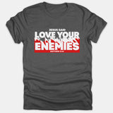 A dark gray t-shirt featuring bold white text that reads 'Jesus Said Love Your Enemies' with 'Matthew 5:44' printed across a distressed red background graphic.
