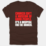 A brown Christian t-shirt featuring bold red and white text that reads, 'Church isn't a museum of good people. It's a hospital for the broken.'