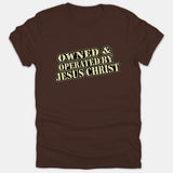 A brown t-shirt featuring a distressed vintage-style typography graphic that reads OWNED & OPERATED BY JESUS CHRIST across the chest.