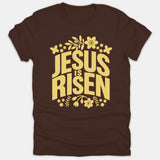 A brown t-shirt featuring the text 'JESUS IS RISEN' in bold yellow typography, framed by decorative flowers and butterflies.