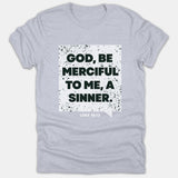 A light gray Christian t-shirt featuring a distressed white box graphic with bold black text reading 'GOD, BE MERCIFUL TO ME, A SINNER.' above Luke 18:13.