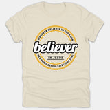 A beige Christian T-shirt featuring a circular badge emblem that reads 'believer in Jesus' surrounded by John 3:36 scripture text.