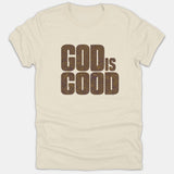 A beige Christian t-shirt featuring the phrase 'GOD IS GOOD' printed in bold block typography filled with a retro wavy purple and gold pattern.