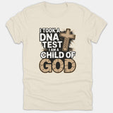 A beige graphic t-shirt with a wooden cross containing a DNA strand design, accompanied by the text 'I took a DNA test I am a child of God'.