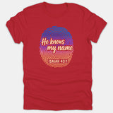 A red t-shirt featuring a colorful gradient fingerprint graphic with the phrase 'He knows my name' and the scripture reference 'ISAIAH 43:1'.