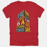 A red t-shirt featuring a 3D tower graphic formed by colorful typography displaying the Bible verse, 'The name of the Lord is a strong tower; the righteous run to it and are safe.'