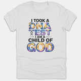 A white Christian T-shirt featuring the graphic text 'I TOOK A DNA TEST I AM A CHILD OF GOD' in black and colorful tie-dye typography.